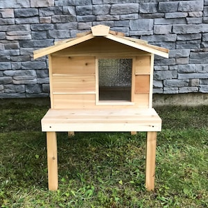 May include: A wooden cat house with a raised platform and a small window. The house is made of natural wood and has a simple design.