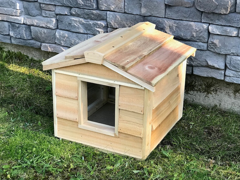 May include: A wooden dog house with a sloped roof and a rectangular entrance. The house is made of natural wood and has a light brown color.