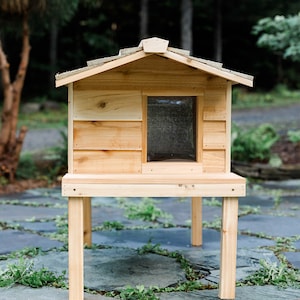 May include: A wooden cat house with a raised platform and a small window. The house is made of natural wood and has a simple design.