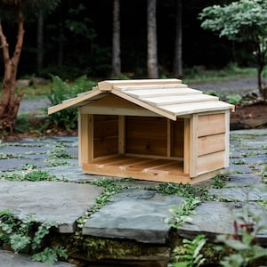 May include: A small, wooden dog house with a sloped roof and open front. The structure is made of light-colored wood and sits on a stone patio with greenery. The dog house is designed to provide shelter for a small pet.