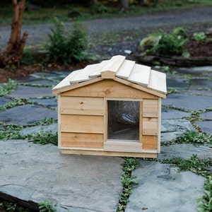 May include: A wooden dog house with a slanted roof and a small window. The house is made of light brown wood and is sitting on a stone patio.