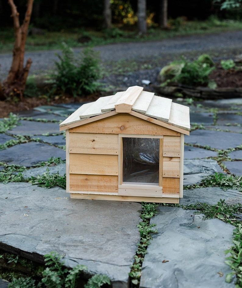 May include: A wooden dog house with a slanted roof and a small window. The house is made of light brown wood and is sitting on a stone patio.