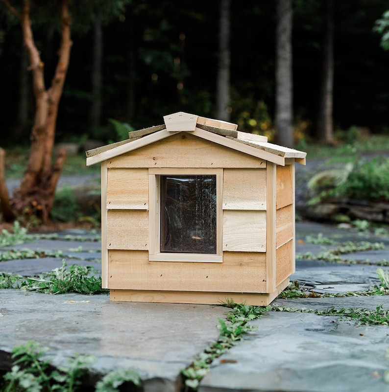May include: A wooden cat house with a small window and a shingled roof. The house is made of natural wood and has a rustic look.
