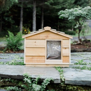 May include: A wooden dog house with a slanted roof and a small window. The house is painted a light brown color and is sitting on a stone patio.