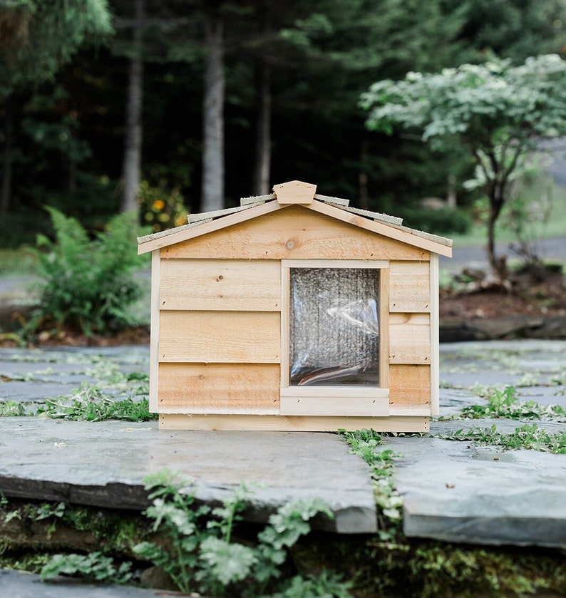 May include: A wooden dog house with a slanted roof and a small window. The house is painted a light brown color and is sitting on a stone patio.