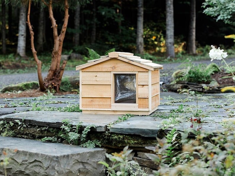 May include: A wooden cat house with a slanted roof and a small window. The house is made of cedar wood and has a natural finish. It is sitting on a stone patio.