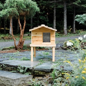 May include: A wooden cat house with a shingled roof, raised on four legs, sits on a stone patio. The house has a small window and is painted a natural wood color.