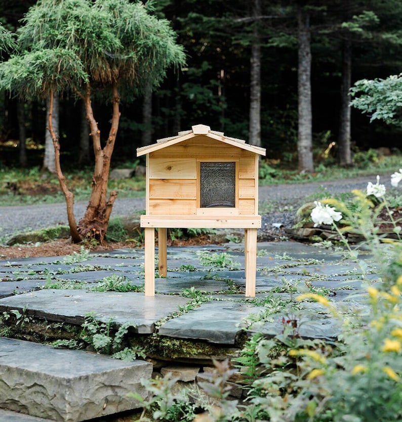 May include: A wooden cat house with a shingled roof, raised on four legs, sits on a stone patio. The house has a small window and is painted a natural wood color.