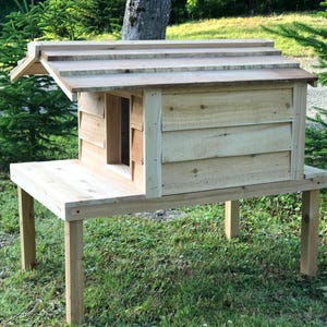 May include: A wooden cat house with a raised platform and a slanted roof. The house is made of natural wood and has a small entrance hole. The house is elevated on four wooden legs.