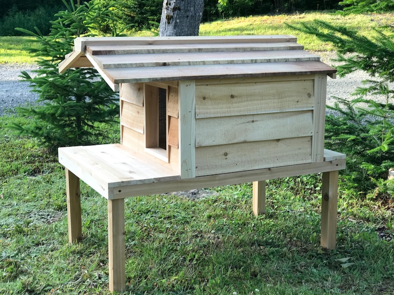 May include: A wooden cat house with a raised platform and a slanted roof. The house is made of natural wood and has a small entrance hole. The house is elevated on four wooden legs.