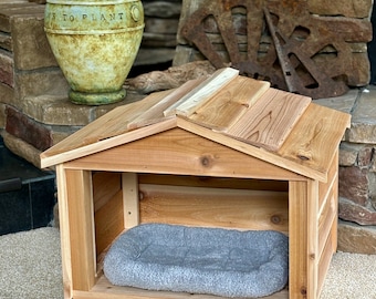 May include: A wooden dog house with a gray dog bed inside. The house has a shingled roof and is made of cedar wood.