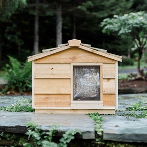 May include: A wooden dog house with a slanted roof and a small window. The house is painted a light brown color and is sitting on a stone patio.