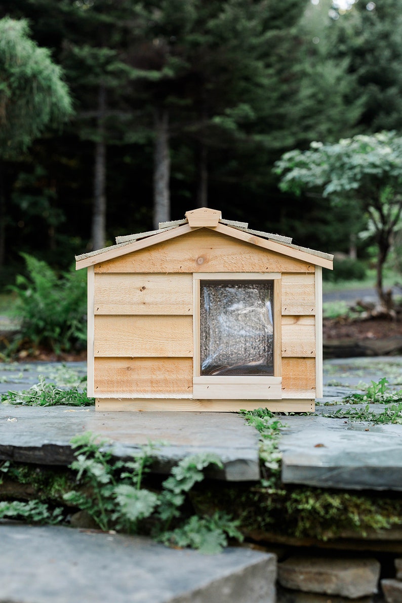 May include: A wooden dog house with a slanted roof and a small window. The house is painted a light brown color and is sitting on a stone patio.