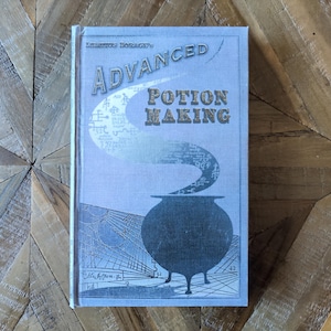 May include: A light blue hardback book titled Libatius Borage's Advanced Potion Making. The cover features a dark blue illustration of a three-legged cauldron with a swirling vapour trail containing various symbols rising above it.