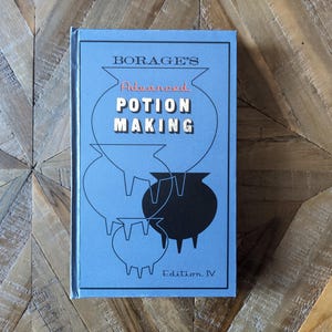 Può includere: Un libro con copertina rigida azzurro intitolato Borage's Advanced Potion Making Edition IV. La copertina presenta illustrazioni a linee nere di calderoni a tre gambe, con un calderone nero pieno e il titolo in lettere bianche e rosse.