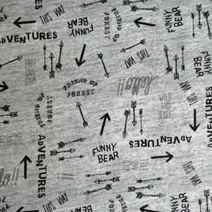 Könnte beinhalten: Grauer Stoff mit einem sich wiederholenden Muster aus schwarzen Pfeilen und Text, der "Adventures", "Funny Bear", "Friend of Forest", "This Way", "Wild and Free" und "Hello!" sagt.
