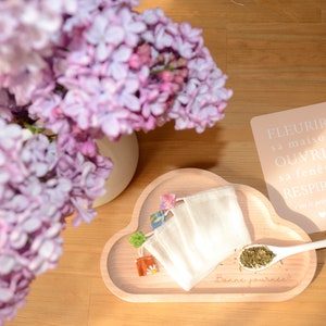 Peut inclure: Un plateau en bois clair en forme de nuage, portant l'inscription "Bonne journée" gravée sur sa surface. Sur le plateau se trouvent trois petites pochettes en tissu blanc avec des languettes à motifs colorés et une cuillère blanche.