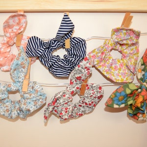 Peut inclure: Six scrunchies en tissu avec différents motifs floraux et un motif rayé. Les scrunchies sont accrochés à un fil métallique avec des pinces à linge en bois.