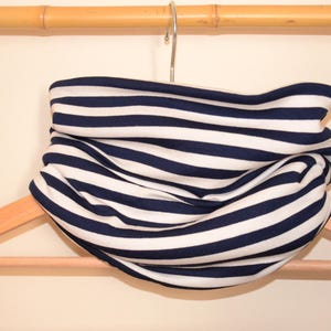 Op de afbeelding: Een witte en marineblauwe gestreepte nekwarmer van stof die aan een houten hanger hangt.