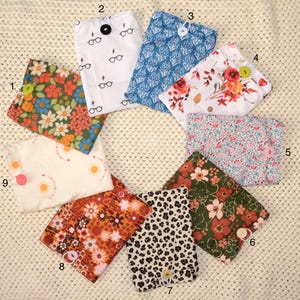 Può includere: Una collezione di nove pochette in tessuto con chiusure a bottone, ognuna con un motivo unico. I disegni includono stampe floreali, geometriche e animalier in vari colori. Una pochette ha un motivo a fulmine e occhiali.