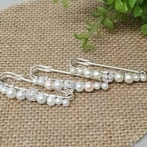 Op de afbeelding: Drie zilverkleurige veiligheidsspelden versierd met imitatieparels en strass. De spelden hebben een gebogen ontwerp met een reeks witte en roze parels en een cluster van strass. De spelden zijn gerangschikt op een getextureerd, neutraal getint oppervlak.