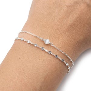 Peut inclure: Deux bracelets argentés délicats sont présentés. L'un comporte une chaîne avec de petits accents scintillants, tandis que l'autre a une petite pierre précieuse blanche et ronde. Les bracelets sont portés sur un poignet.