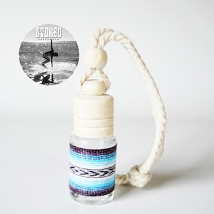 May include: A clear glass bottle with a blue and white patterned label. The bottle has a wooden stopper and a white rope attached to it. The bottle is a car air freshener.