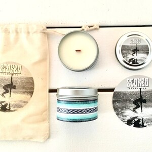 May include: A set of surf-themed products including a candle, a sticker, a bag, and a tin. The candle is in a silver tin with a wooden wick. The sticker is round and features a surfer riding a wave. The bag is made of natural fabric and has a surfer graphic. The tin is silver with a blue and white striped design. The text "STOKED" is featured on the bag, sticker, and tin.