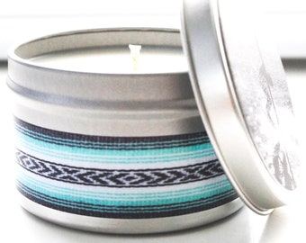 May include: A silver candle tin with a teal, black, and white striped design. The candle is white and has a single wick.
