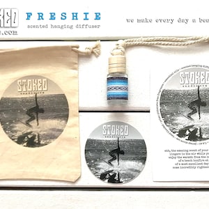 May include: A white drawstring bag with a black and white image of a surfer on a wave. The bag is labeled "STORED candle.com". A small glass bottle with a blue and white patterned label is hanging from a white cord. The bottle is labeled "STORED". A white card with black text reads "FRESHIE scented hanging diffuser we make every day a beach day".