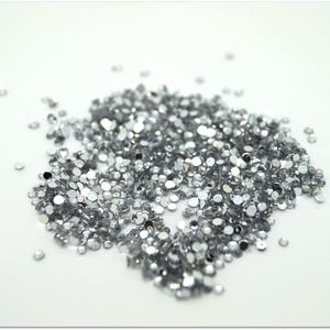 Può includere: Un mucchio di piccole strass argentati sparsi su una superficie bianca. Gli strass sono rotondi e sfaccettati, riflettendo la luce. Variano leggermente di dimensioni e sono raggruppati, con alcuni sparsi sui bordi.