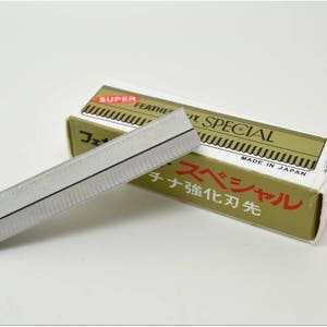 Peut inclure: Une lame de rasoir argentée avec une bande noire au centre, à côté d'une boîte dorée et rouge qui dit "Super Feather Special" et "Fabriqué au Japon".