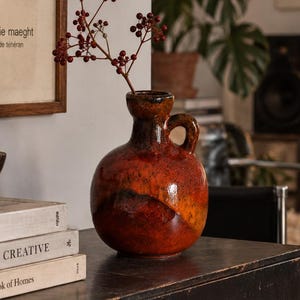 May include: A round, glazed ceramic jug with a single handle, featuring a gradient of deep red, orange, and brown tones. A thin, woody stem with small dark red berries is placed inside the jug. The jug sits on a dark, weathered wooden surface.