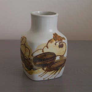 Può includere: Un vaso in ceramica bianca con base quadrata e collo stretto. Il vaso è decorato con un motivo astratto marrone e arancione.