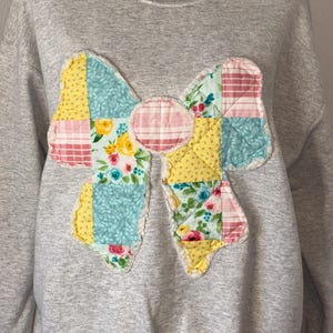May include: A gray sweatshirt featuring a large, patchwork bow applique. The bow is made of various patterned fabrics, including floral, plaid, and solid colors in yellow, blue, and pink. The applique has a scalloped edge.