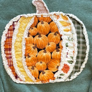 May include: A sage green sweatshirt featuring a large, central pumpkin-shaped applique. The pumpkin is constructed from various fabric panels in plaid, floral, and pumpkin-print patterns, all finished with frayed, raw-edge white seams.