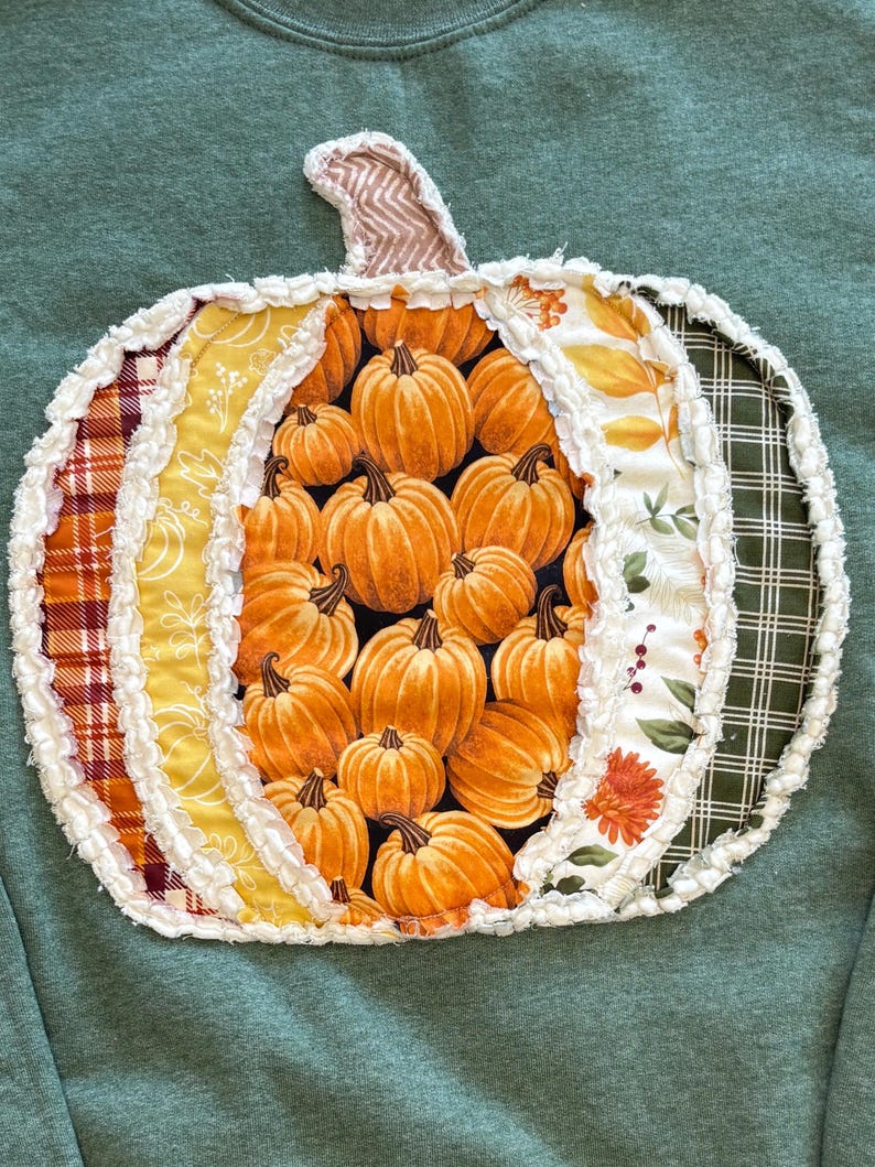 May include: A sage green sweatshirt featuring a large, central pumpkin-shaped applique. The pumpkin is constructed from various fabric panels in plaid, floral, and pumpkin-print patterns, all finished with frayed, raw-edge white seams.