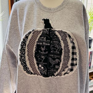 May include: A light gray crewneck sweatshirt featuring a large, central pumpkin applique. The pumpkin is constructed from various black, white, and gray fabric patches with frayed edges, displaying farm-themed graphics and text like Farm House.