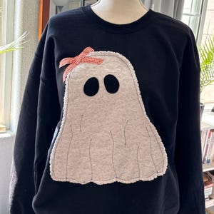 May include: A black long-sleeve sweatshirt featuring a large, white, textured ghost appliqué on the front. The ghost has two black oval eyes and a small orange and white checkered bow attached to its upper left side, with visible stitching.