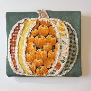May include: A square dark green fabric pillow featuring a large, central pumpkin-shaped applique. The pumpkin is constructed from multiple vertical fabric panels in various patterns, including orange pumpkin prints, plaid, and floral motifs.