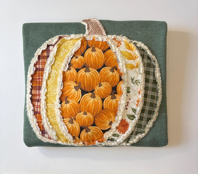 May include: A square dark green fabric pillow featuring a large, central pumpkin-shaped applique. The pumpkin is constructed from multiple vertical fabric panels in various patterns, including orange pumpkin prints, plaid, and floral motifs.