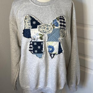 May include: A heather gray crewneck sweatshirt featuring a large, central bow-shaped appliqué. The bow is constructed from a patchwork of various blue and white fabrics, including floral, gingham, and polka dot patterns with frayed edges.