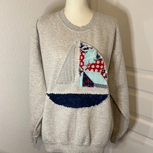 May include: A light gray sweatshirt with a sailboat applique. The sailboat design features a striped sail, a red and white patterned sail, and a dark blue base. The sweatshirt has a crew neck and long sleeves.