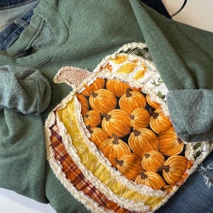 May include: A sage green sweatshirt featuring a large, central pumpkin-shaped appliqu&eacute;. The pumpkin is constructed from multiple fabric patches in shades of orange, yellow, and plaid, with frayed, raw-edge seams and a chevron-patterned stem.