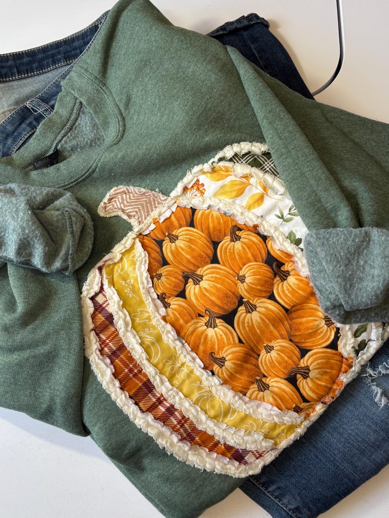 May include: A sage green sweatshirt featuring a large, central pumpkin-shaped appliqu&eacute;. The pumpkin is constructed from multiple fabric patches in shades of orange, yellow, and plaid, with frayed, raw-edge seams and a chevron-patterned stem.