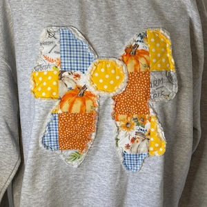 May include: A light gray crewneck sweatshirt featuring a large, central bow-shaped appliqué made from various quilted fabric patches. The patches include patterns of orange pumpkins, yellow polka dots, blue gingham, and text reading FRESH and FARM.