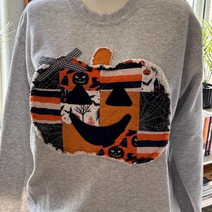 May include: A light gray crewneck sweatshirt featuring a large, quilted pumpkin appliqué on the chest. The pumpkin is constructed from various fabric patches in orange, black, and white, displaying patterns of spiderwebs, bats, and ghosts.