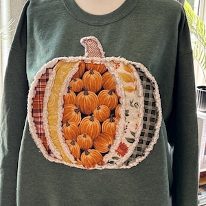 May include: A dark green crewneck sweatshirt featuring a large, central pumpkin-shaped appliqué. The pumpkin is constructed from various fabric panels in orange, yellow, and plaid patterns, with frayed, raw-edge seams and a textured stem.