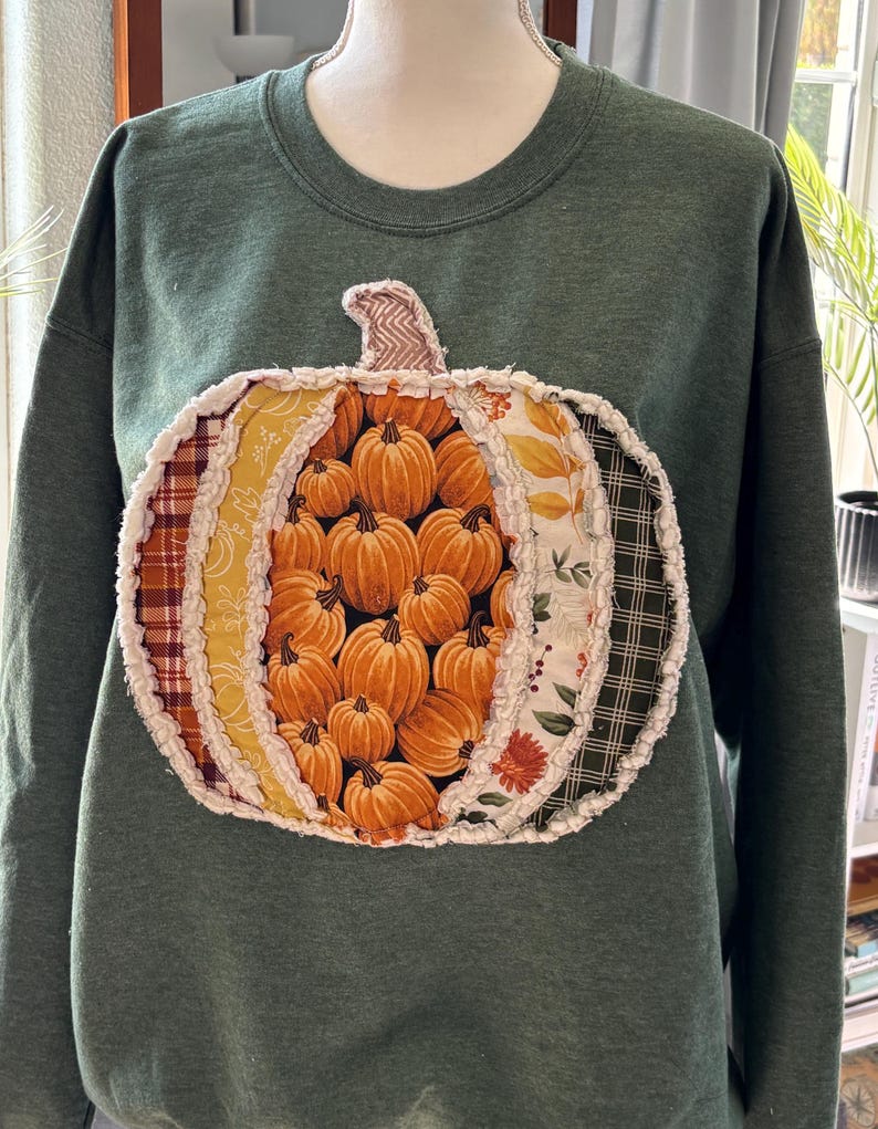 May include: A dark green crewneck sweatshirt featuring a large, central pumpkin-shaped appliqu&eacute;. The pumpkin is constructed from various fabric panels in orange, yellow, and plaid patterns, with frayed, raw-edge seams and a textured stem.