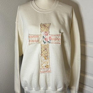 May include: Cream-colored sweatshirt featuring a cross applique. The cross is made of floral and striped fabric patches in shades of pink, yellow, and cream. The sweatshirt has a crew neck and long sleeves, suitable for casual wear.